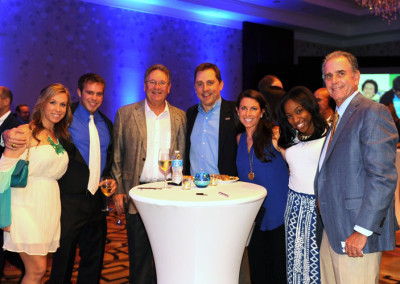 Group of seven adults smiling at a social event with drinks, standing around a high-top table in a room with blue lighting.