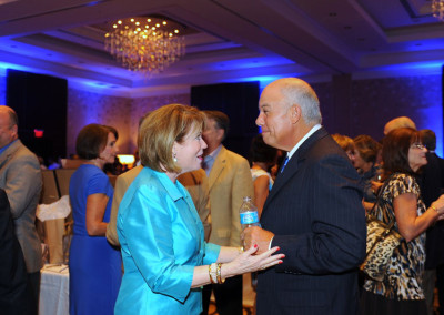 Two older adults engaging in a conversation at a formal event with other attendees in the background.