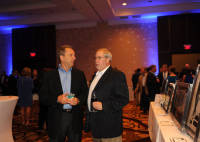Two men conversing while holding drinks at a business event in a banquet hall with attendees in the background.