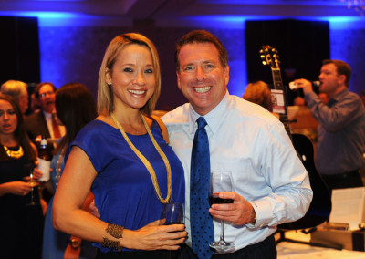 A man and a woman, both smiling and holding glasses of wine, posing together at a formal event.