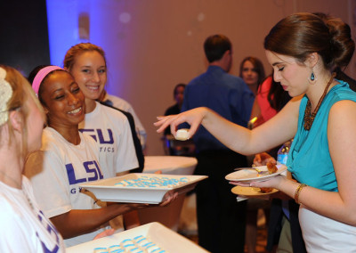 A woman offers cupcakes on a tray to attendees at an indoor event, with others in athletic wear smiling in the background.