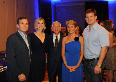 Five adults smiling at a formal event, three men and two women, dressed in business and evening attire, standing under dim lighting with a blue background.
