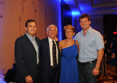 Four people posing for a photo at an event, with two men in suits and one man in a casual shirt alongside a woman in a blue dress.