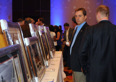 A man in a suit examining framed pictures at an art auction event, with other attendees in background.