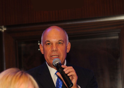 A bald man in a suit speaking into a microphone in a dimly lit room.