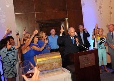 A man gives a toast at a formal event while a group of people hold up their glasses in celebration.