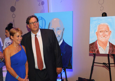A woman in a blue dress and a man in a suit stand next to each other, with paintings on easels behind them at an event.