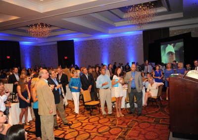 Crowd of people watching a presentation at a formal event in a large room with chandeliers and a projector screen.