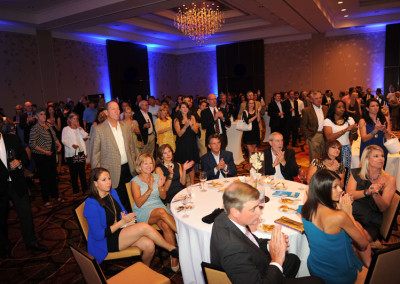 Audience in a formal banquet hall clapping and watching a presentation, with some seated at tables and others standing.