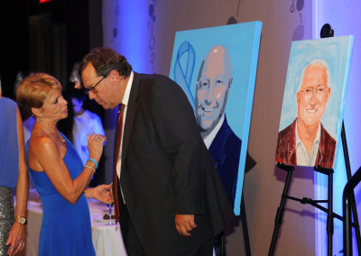 A man and a woman in conversation at an event, with two portrait paintings on easels in the background.