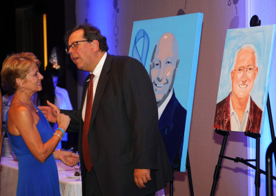 A man in a suit and a woman in a blue dress converse at an event, with two portrait paintings on easels in the background.