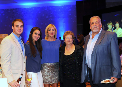 Five people posing together at a social event, three women and two men, with a blue-lit background and event attendees visible on a screen behind.