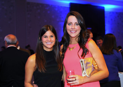 Two women smiling at a formal event, one holding a glass of wine, with others in the background and blue lighting.