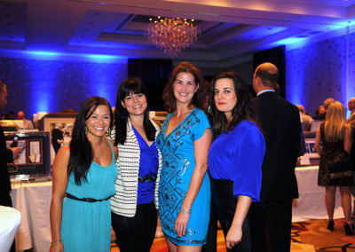Four women smiling at a gala event in a room with blue lighting and a chandelier, with other attendees in the background.