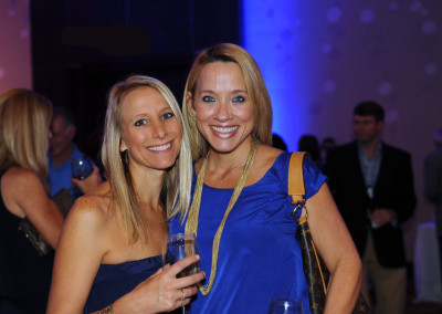 Two women smiling at a social event, holding drinks, with a blurred party background.