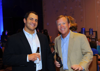 Two smiling men holding beer bottles at a conference event. one man wears a black jacket and checked shirt, the other a yellow jacket and blue shirt.