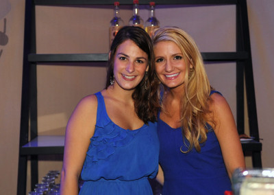 Two women in blue dresses smiling at a bar event, standing in front of a shelf with bottles.