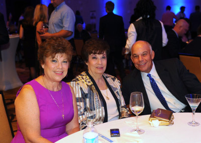 Three senior adults, two women and a man, smiling and sitting at a table during a gala event, with drinks and a blue-lit background.