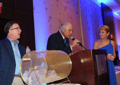Three people at a gala; a man speaks at a podium while another man and a woman in a blue dress stand beside him, with a raffle drum on the table.
