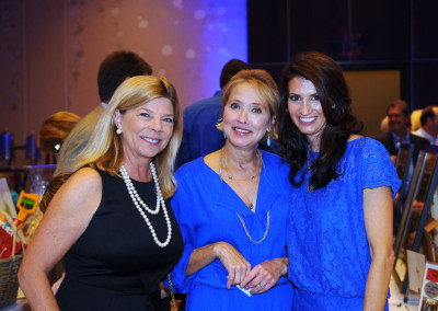 Three women smiling at a gala event, dressed in semi-formal attire, with a blurred background of guests and lights.