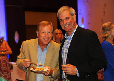 Two smiling men at a social event, one holding a plate of food and the other a wine glass.