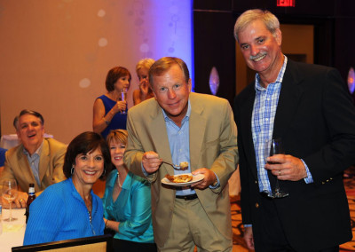 Three smiling people at a social event, two standing with one holding a wine glass, and one seated eating dessert.