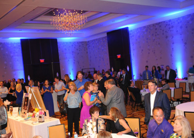 Guests at a formal indoor event mingle and converse under a large chandelier in a ballroom with blue ambient lighting.