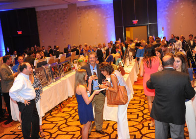 People mingling and viewing auction items at a formal charity event in a banquet hall with blue lighting.