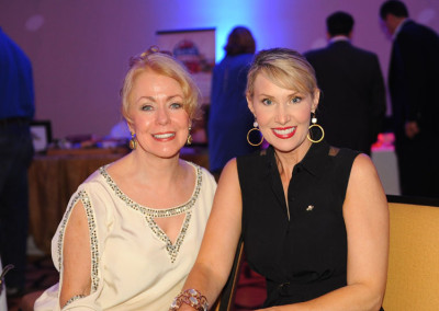 Two women smiling at a social event, one with blonde hair in a white dress, the other in a black dress with large gold earrings.