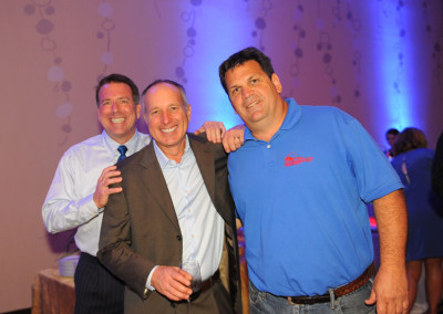Three men smiling at a social event, one in a suit, another in a shirt and tie, and the third in a blue polo shirt.
