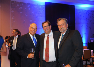 Three men smiling at a formal event, one holding a wine glass, with a blue-lit background and other guests.