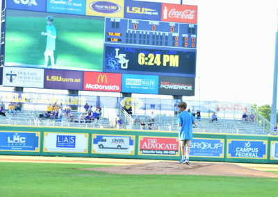 A young boy in a blue shirt stands on a baseball field, looking towards a large scoreboard displaying the time 6:24 pm.