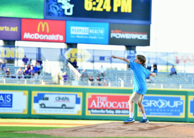 A young boy in a blue shirt throwing the first pitch at a baseball game, with a digital scoreboard in the background.