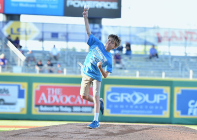 A young boy throws the first pitch at a baseball game, wearing a blue t-shirt and shorts, on a sunny day at a stadium.