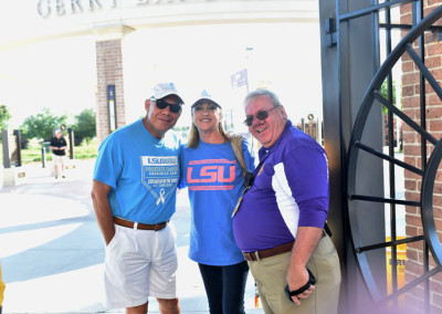 Three people wearing lsu shirts smiling in front of a building with a sign that reads "bertman drive".