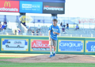 A young boy in a blue shirt and shorts standing on a baseball mound in a stadium, with advertising signs in the background.