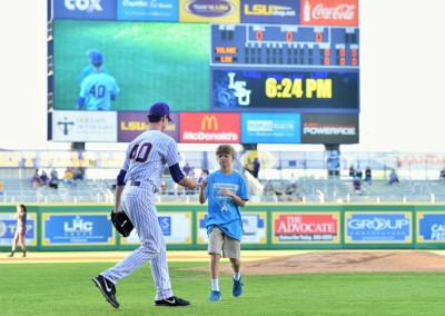 A baseball player in a purple uniform high-fiving a young boy on a baseball field with a large scoreboard in the background.