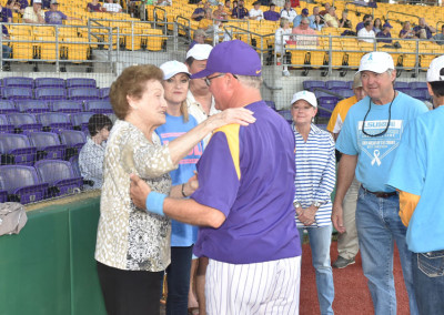 An elderly woman and a baseball coach in purple and gold engage in conversation on a field, with spectators in the background.