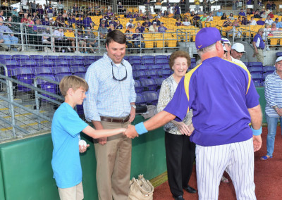 A young boy shakes hands with a baseball coach on the field while a man and an elderly woman watch, with spectators in the stands behind them.