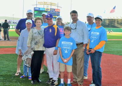 Group of nine individuals, mixed ages and genders, standing on a baseball field in front of a scoreboard. some wear casual attire, others in sports uniforms.