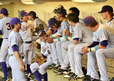 Baseball players in purple uniforms sitting and standing in a dugout, interacting and preparing for a game.