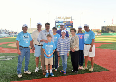 Group of adults and one child in matching blue shirts standing on a baseball field, posing for a photo during an event.