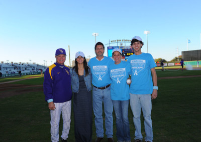 Group of five people wearing blue shirts with ribbons, standing on a baseball field, posing for a photo.
