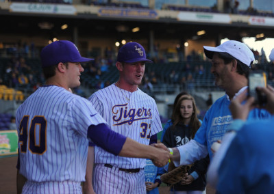 Two baseball players in lsu tigers uniforms shaking hands with a fan on a baseball field.
