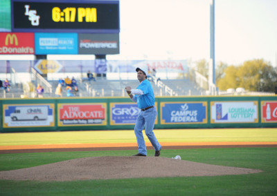 Man pitching a baseball on a well-maintained field with digital scoreboard in background displaying time "6:17 pm".