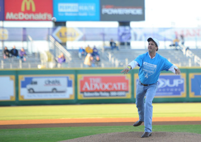 Man in a blue shirt throws the first pitch at a baseball game with stadium advertising banners in the background.