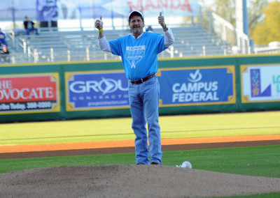 A man in a blue shirt and cap, giving thumbs up, standing on a baseball pitcher's mound in a stadium.