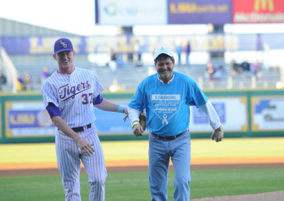 Two men smiling on a baseball field; one in lsu tigers uniform and another in a blue shirt and cap, holding hands in a congratulatory gesture.