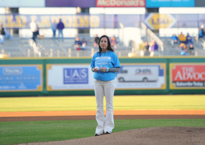 A woman stands on a baseball field holding a microphone, wearing a blue jacket and beige pants, with stadium seats and advertisements in the background.