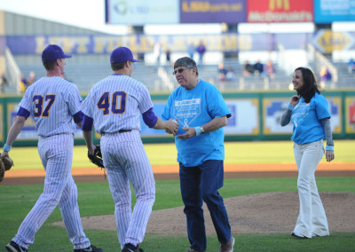 Two baseball players in purple and white uniforms greet a man on a field, while a woman in a blue shirt smiles and walks nearby.